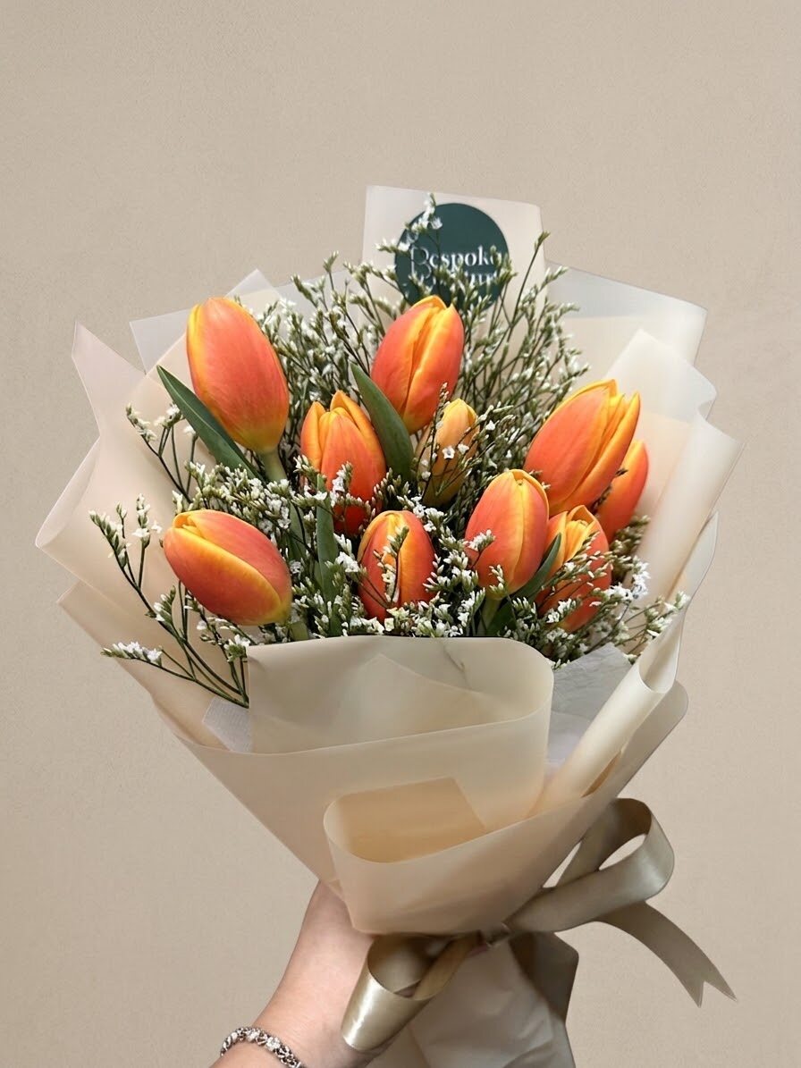 A hand-tied bouquet of orange and yellow tulips with seasonal filler, wrapped in cream paper.