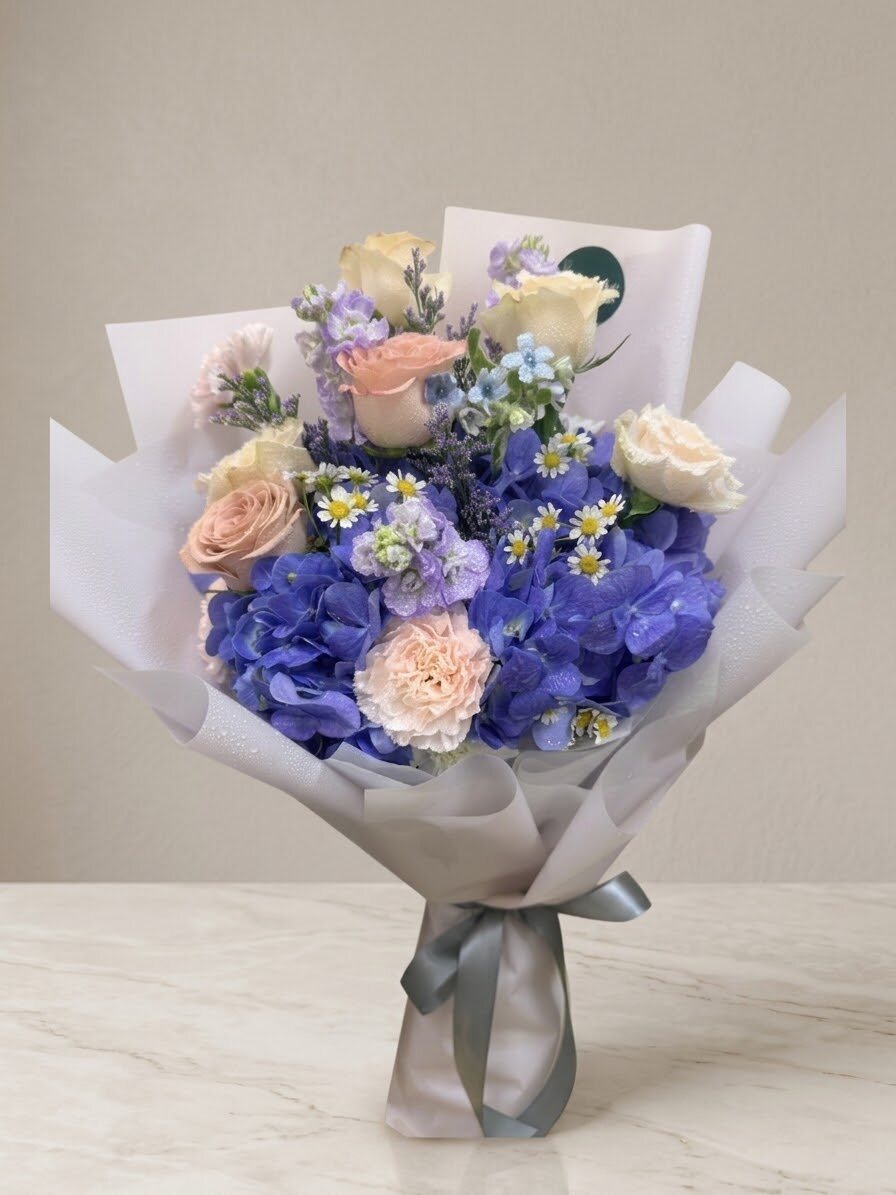 A hand-tied bouquet of blue hydrangea with cream roses, a peach carnation and white chamomile, wrapped in white paper with a grey ribbon.