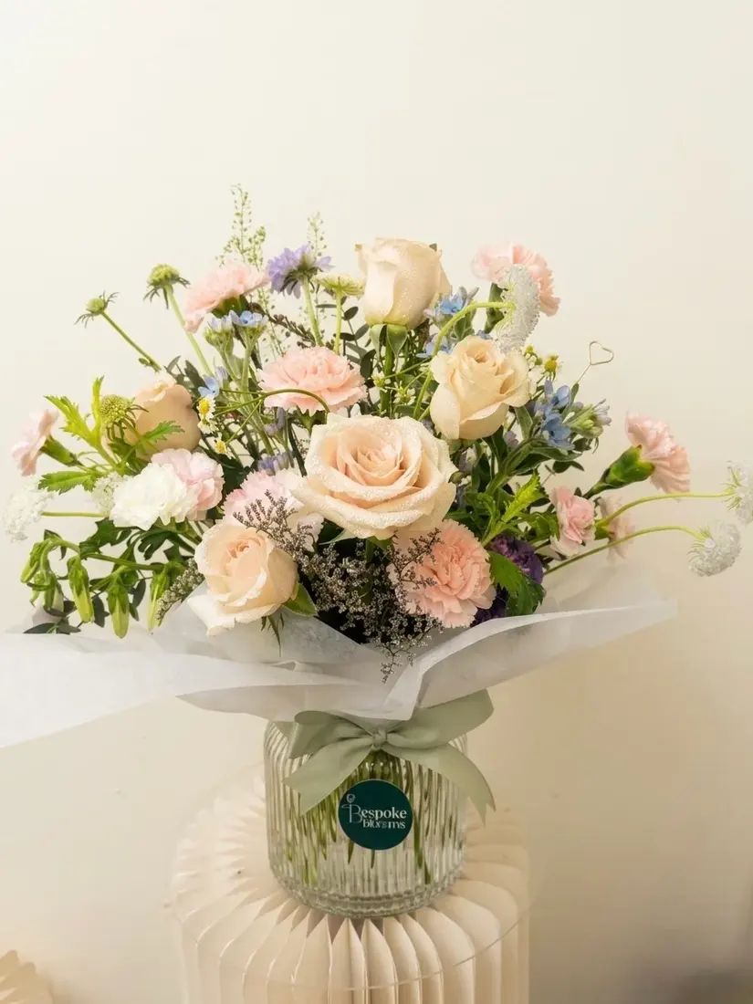 A ribbed glass vase of champagne roses, blush carnations, blue delphinium and scabiosa with a sage ribbon.