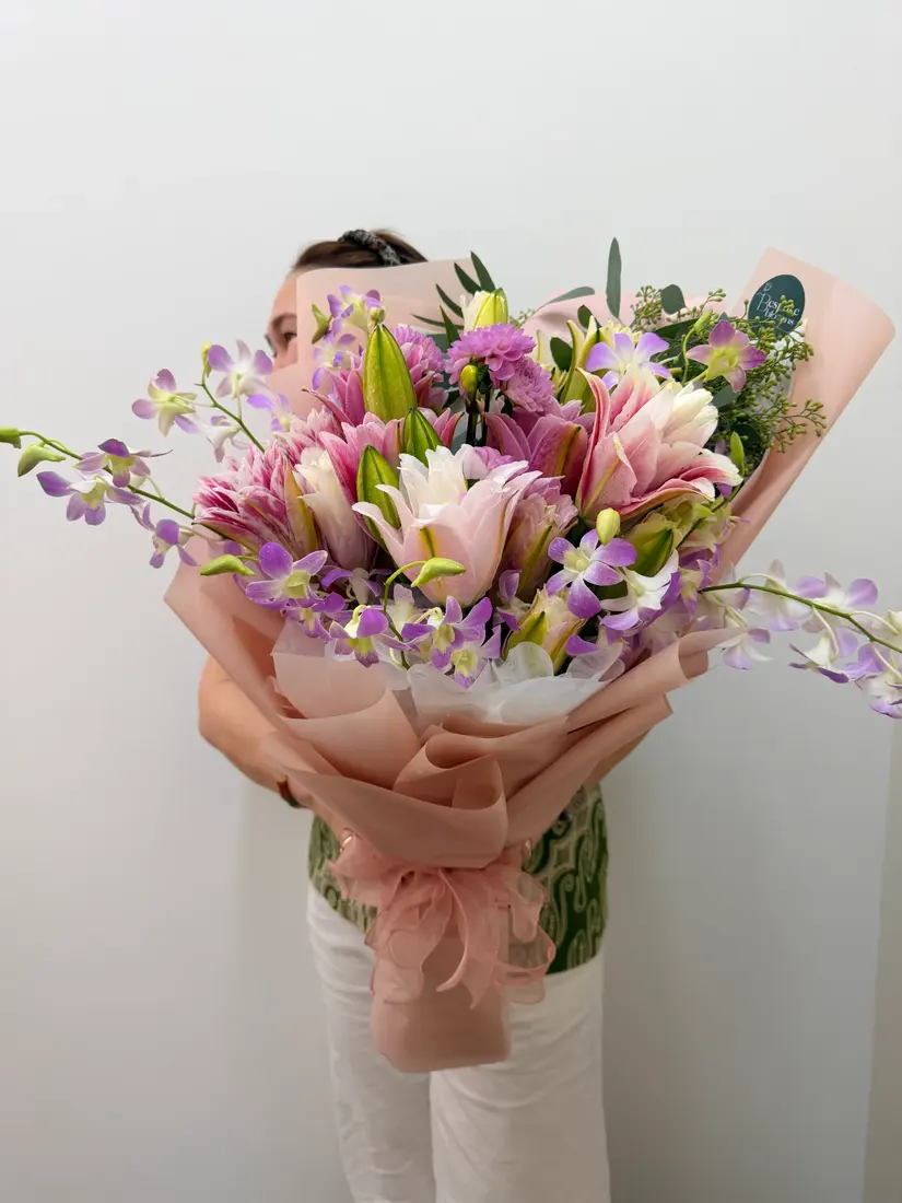 A hand-wrapped bouquet of pink oriental lilies and purple orchids in blush paper with a chiffon bow.
