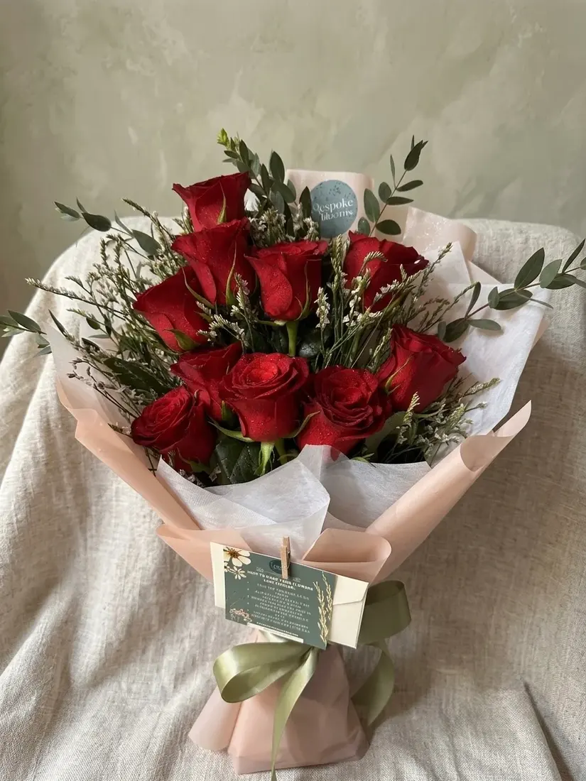 A hand-wrapped bouquet of deep red roses with eucalyptus and white statice, tied with an olive ribbon.