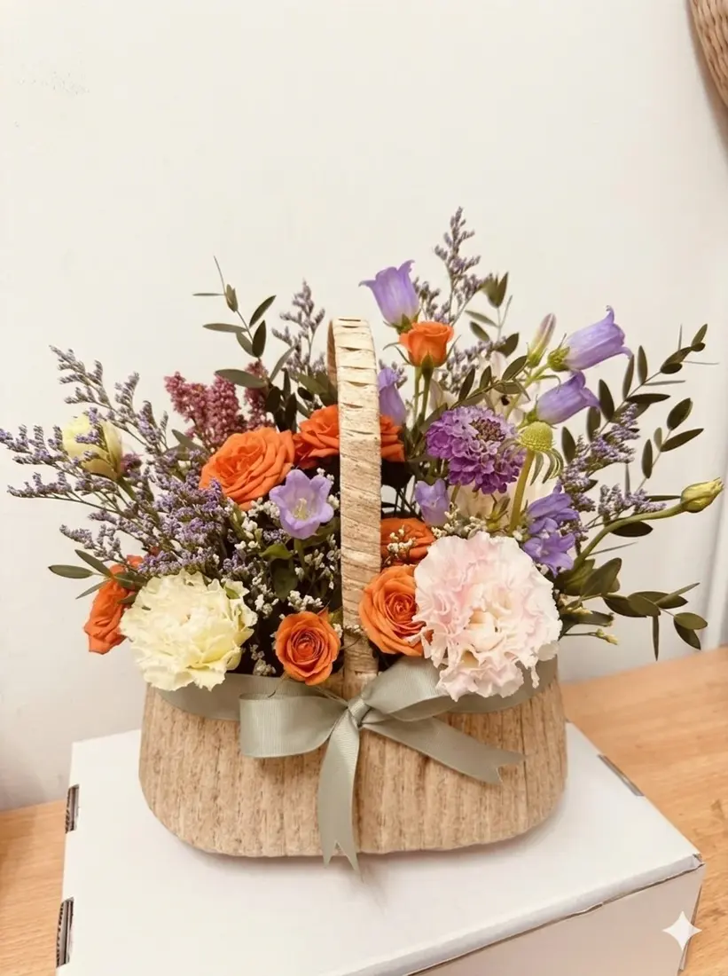 A woven basket of orange spray roses, blush carnations, purple campanula and eucalyptus, finished with a sage ribbon.
