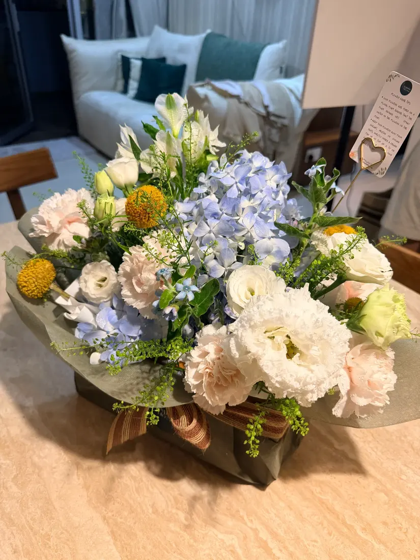 A gathered arrangement of blue hydrangea, white lisianthus, blush carnations and yellow craspedia wrapped in sage paper.