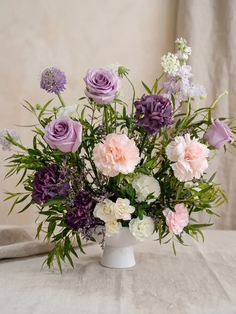 A white footed vase of lavender roses, plum and blush carnations, white hydrangea and scabiosa on a linen-draped table.